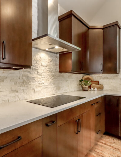 Modern kitchen with dark wood cabinets, white stone backsplash, and a smooth countertop. Features a built-in cooktop and a range hood beneath large windows with greenery outside.