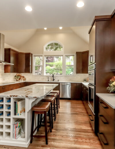 Modern kitchen with dark wood cabinets, stainless steel appliances, and a central island with barstools. A decorative plant sits on the counter, and large windows provide natural light.