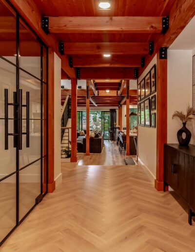 Modern hallway with wooden ceiling beams, glass doors on the left, herringbone wood floor, and a black console with decor on the right, leading to an open living and dining area.