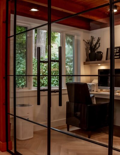 A modern home office with a glass wall, built-in desk, computer, books, chair, and large window overlooking greenery.
