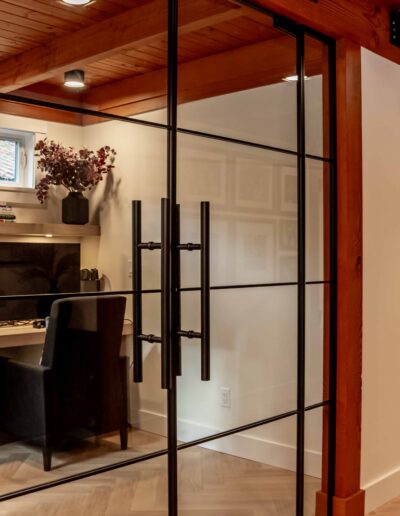 A modern home office with a desk, computer, and chair, enclosed by glass doors, is adjacent to a staircase and wooden beams in a contemporary interior.