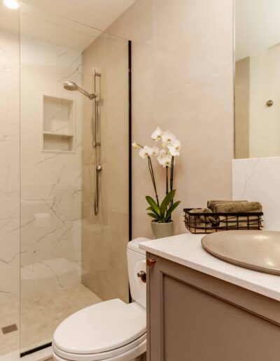 Modern bathroom with a walk-in glass shower, marble walls, a round vessel sink, wall-mounted faucets, and a potted orchid on the vanity.