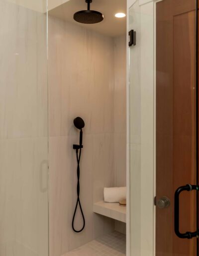 Modern walk-in shower with white tile walls, a black rainfall showerhead, handheld shower, glass door, and a built-in bench holding folded white towels.