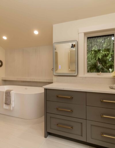 Modern bathroom with a freestanding bathtub, black floor-mounted faucet, large gray vanity, and a window overlooking greenery. Neutral tones and minimal decor create a clean look.