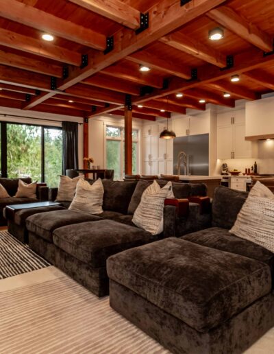 A modern open-concept living room and kitchen with exposed wooden ceiling beams, dark sectional sofas, large windows, and pendant lighting over a kitchen island.