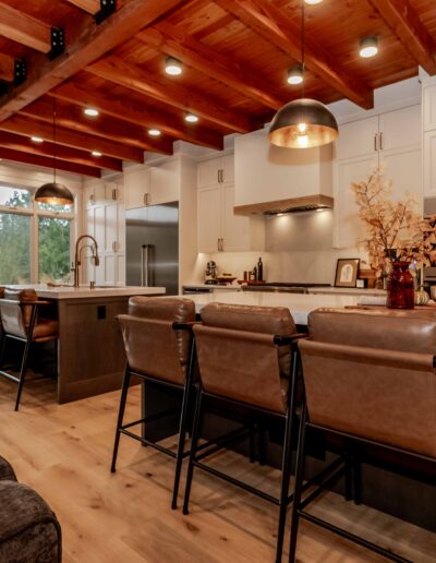 Modern kitchen with wooden beams, white cabinets, stainless steel appliances, large island with bar stools, pendant lighting, and a cozy adjacent seating area.