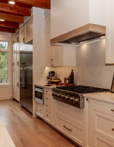 Modern kitchen with white cabinets, stainless steel appliances, wood floor, exposed ceiling beams, and a large window providing natural light.