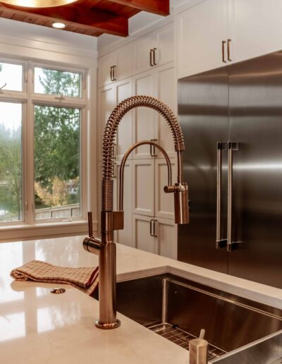 Modern kitchen with a stainless steel double sink, pull-down faucet, white countertops, large refrigerator, and white cabinetry. A window provides a view of trees outside.