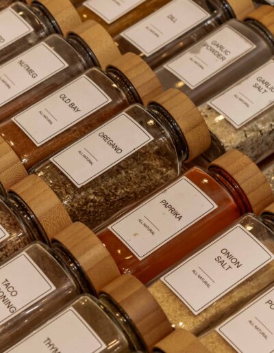 Glass jars with wooden lids, neatly arranged in rows, containing various labeled spices and seasonings such as paprika, oregano, garlic salt, and onion powder.