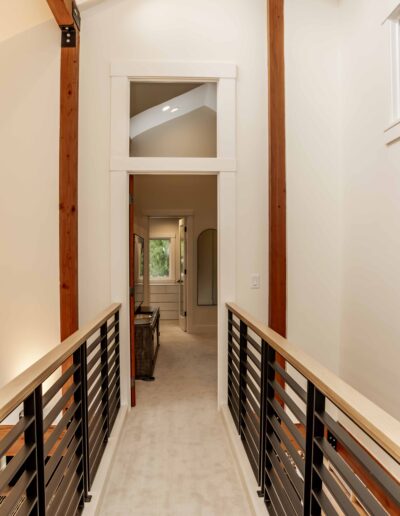 A hallway with beige carpet, wooden beams, and metal railings leads to a room with a large window at the end.