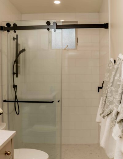 Modern bathroom with a glass-enclosed shower, black fixtures, a small frosted window, white tiles, and white towels hanging on the wall.