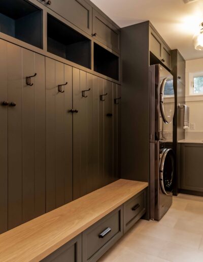 Modern laundry and mudroom with built-in bench, storage cabinets, wall hooks, stacked washer and dryer, and a sink under double windows.