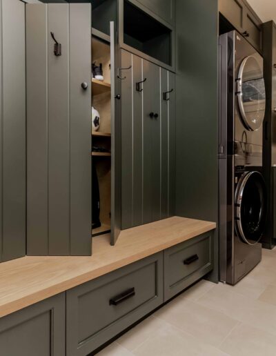 Modern laundry room with green built-in cabinets, open storage lockers, wood bench, stacked washer and dryer, sink, and small window.